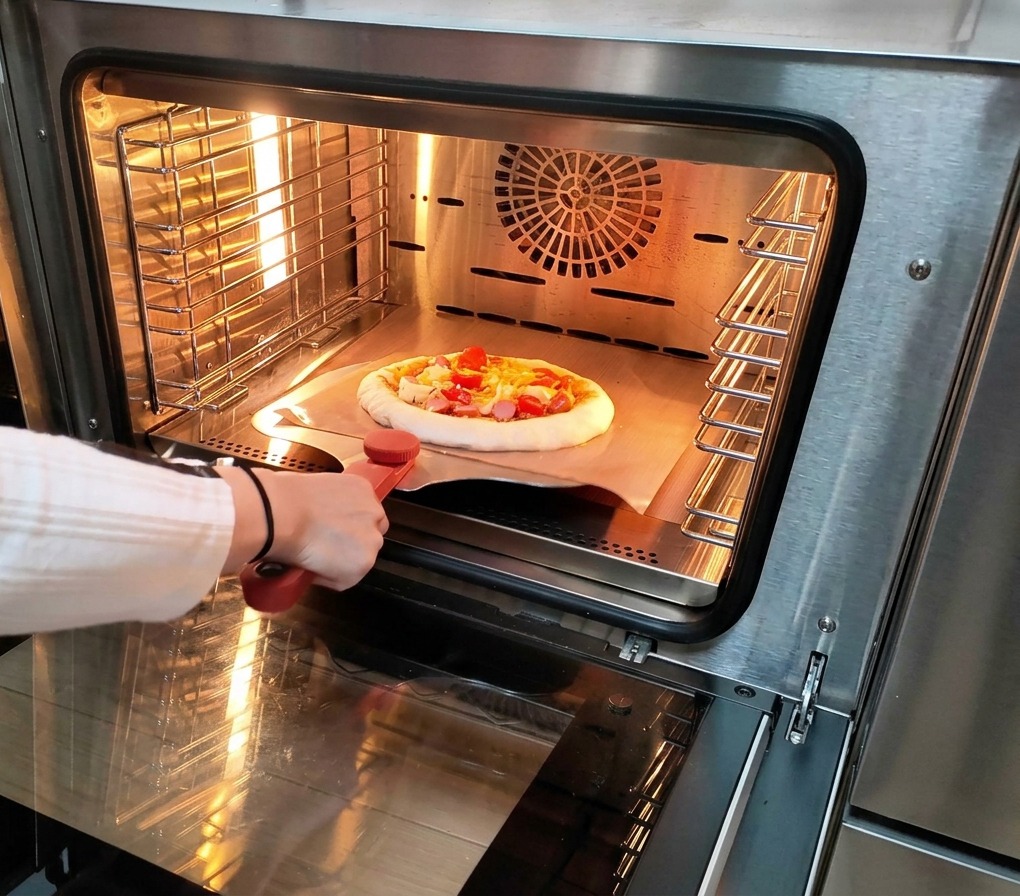 A person using a metal pizza peel to slide a raw pizza topped with sauce, cheese, and meats into a hot stainless steel electric oven.