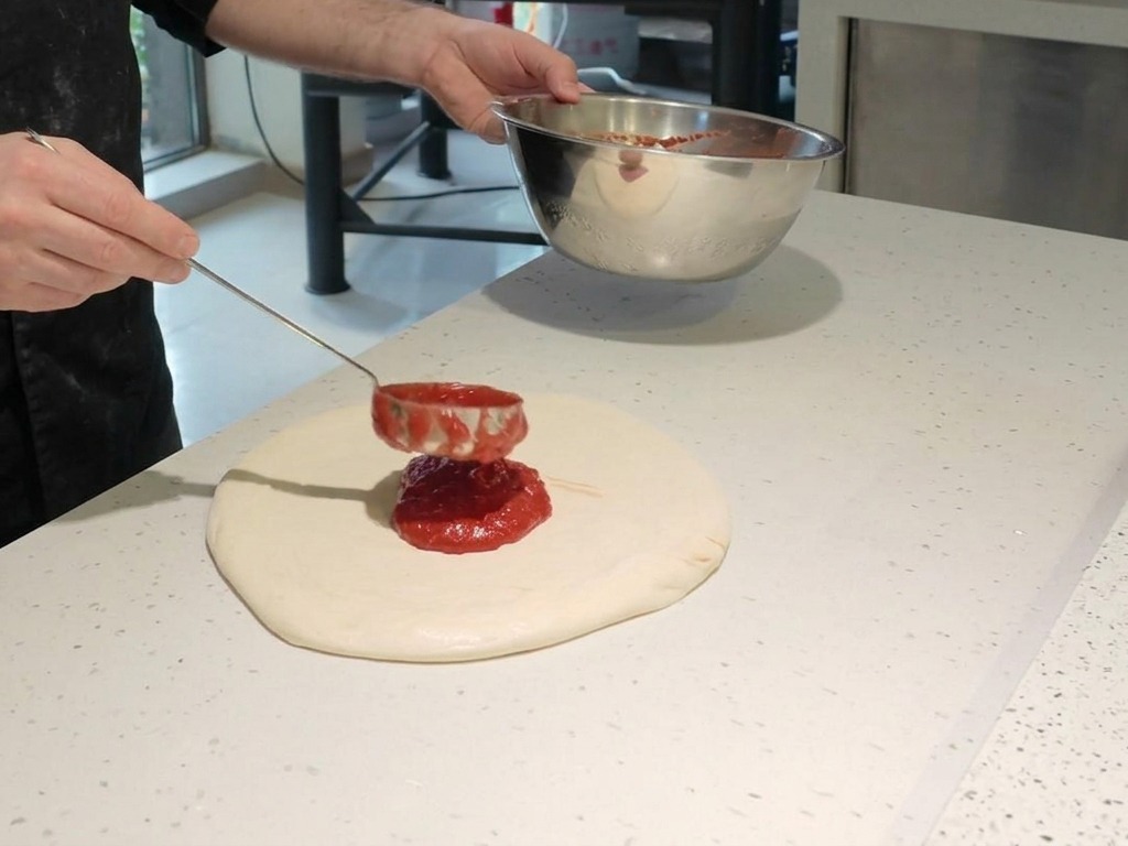 A chef using a metal ladle to spread vibrant red tomato sauce in a spiral pattern over raw pizza dough on a white countertop.