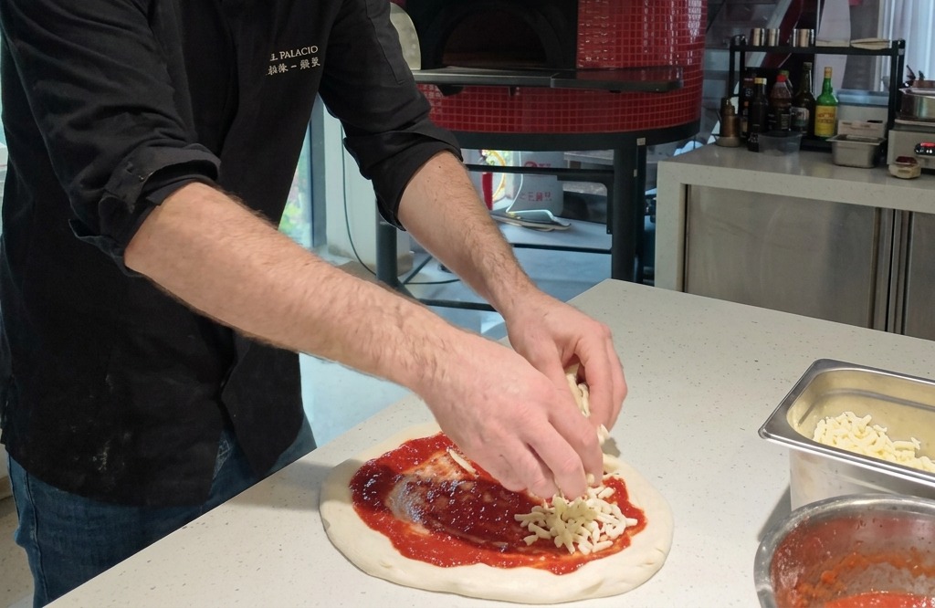 Hands placing chunks of white fresh mozzarella cheese onto a circular pizza base coated with vibrant red tomato sauce.