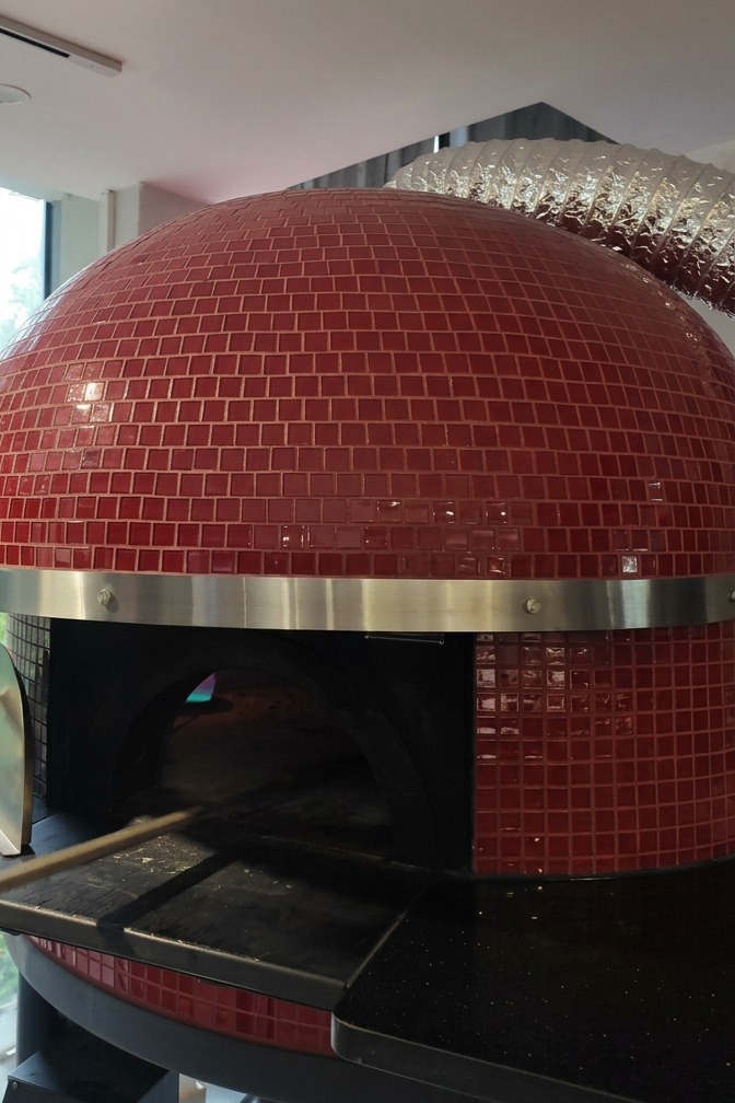 A wood-fired oven with a red-tiled exterior as a pizza is slid inside on a long metal peel to bake.