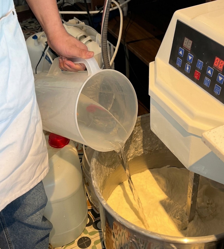Water being poured from a translucent plastic pitcher into a mixer bowl already containing white flour.
