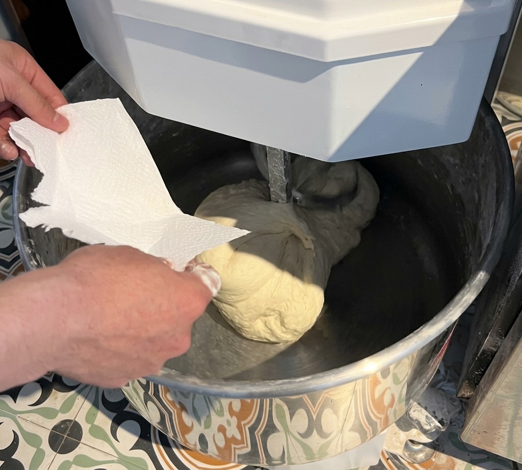 A hand adding salt from a white paper into a mixer bowl containing a large, smooth ball of pizza dough.