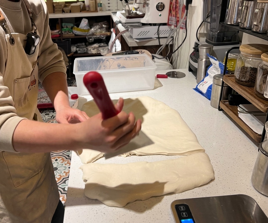 A person using a red-handled dough scraper to cut and weigh portions of pizza dough on a digital scale.