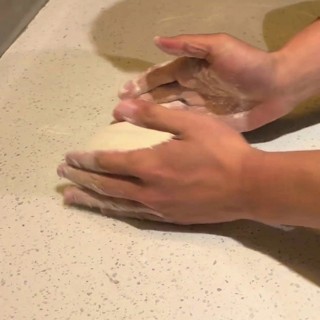 Two hands pressing down on the center of a round piece of fermented pizza dough on a floured white countertop.