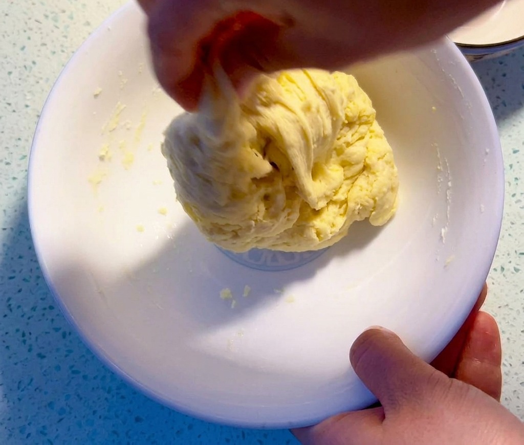A hand pulling and folding a pale, hydrated pizza dough ball inside a white ceramic bowl to develop its structure.