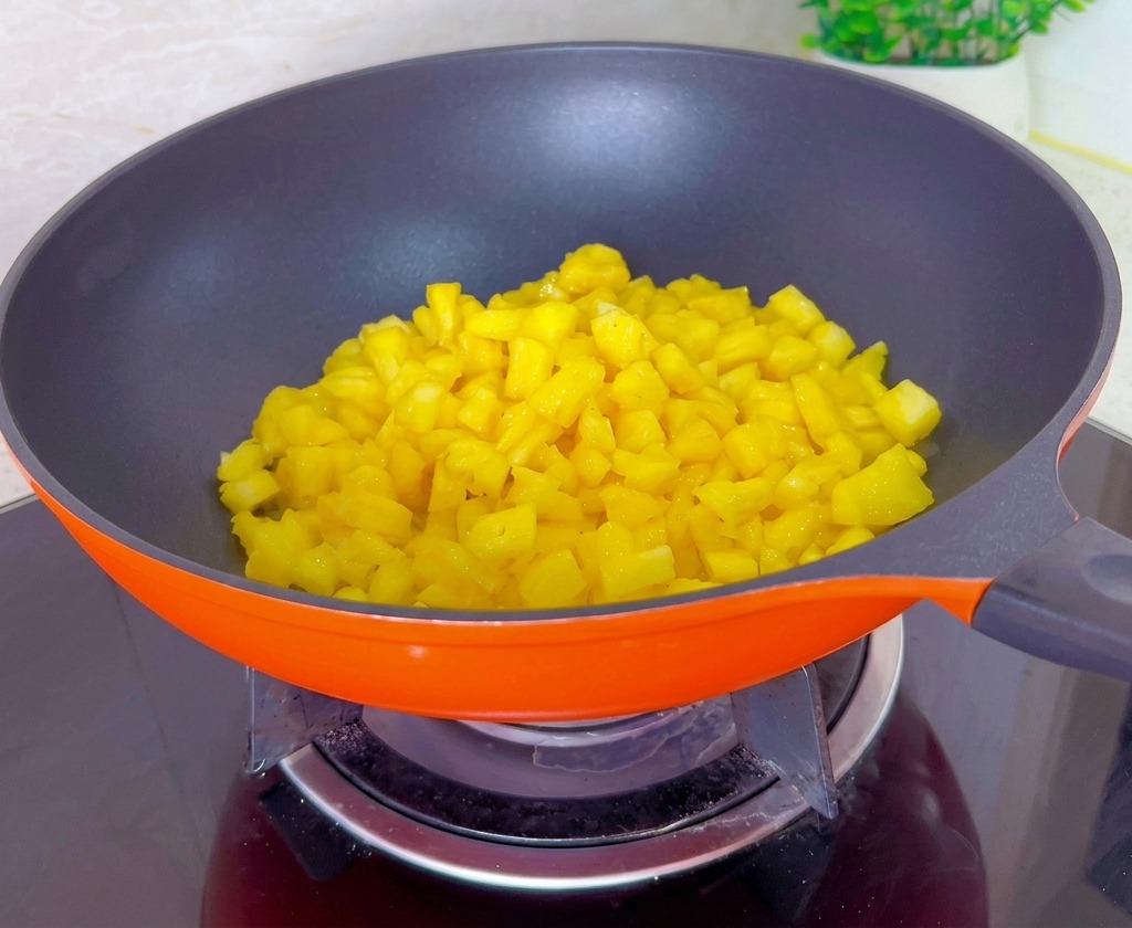 Diced yellow pineapple pieces cooking and reducing in a vibrant orange pan on a glass stovetop.