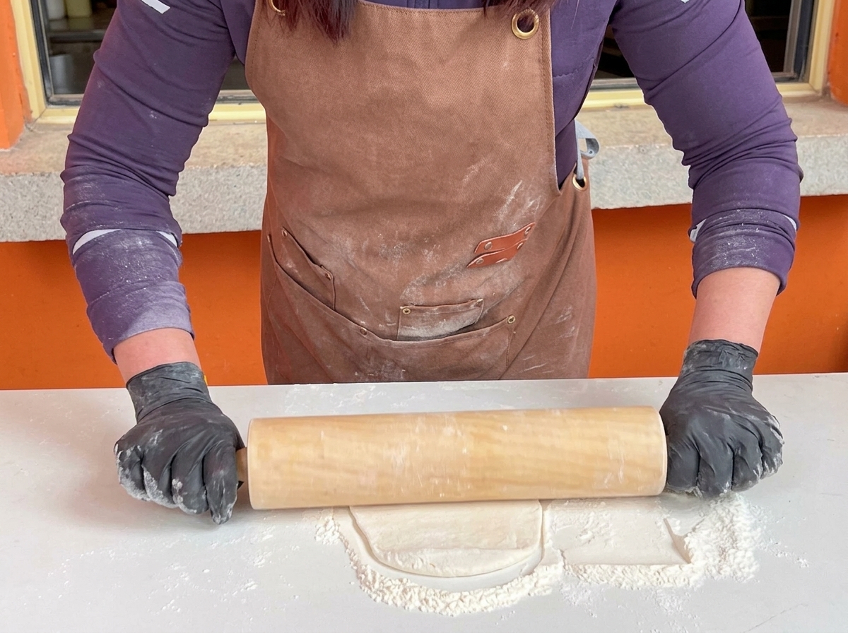 A person in black gloves using a large wooden rolling pin to flatten a pizza dough base on a white floured table.