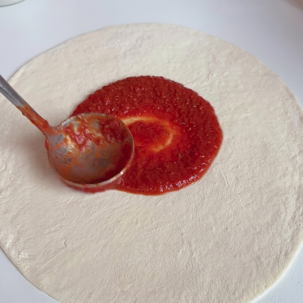 A metal ladle spreading vibrant red tomato sauce in a circular pattern over a raw white pizza dough base.