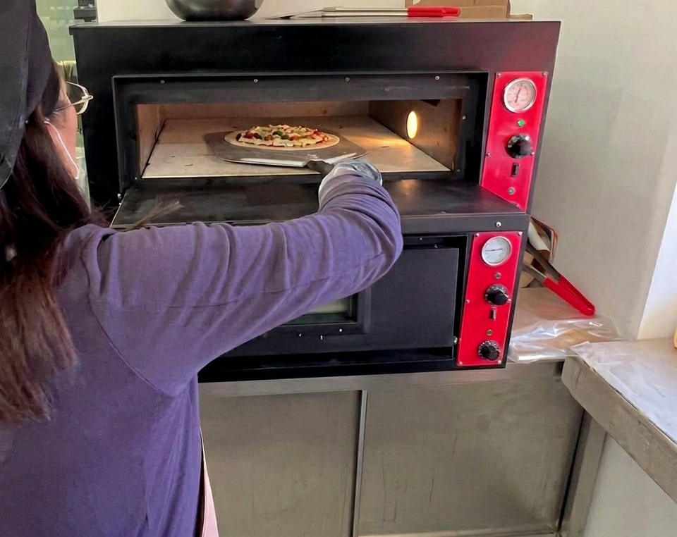 A cook using a long metal peel to place the raw Margherita pizza into a hot professional deck oven.