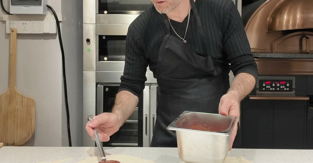 A person using a metal ladle to spread red tomato sauce in a circular motion over a round pizza dough base.