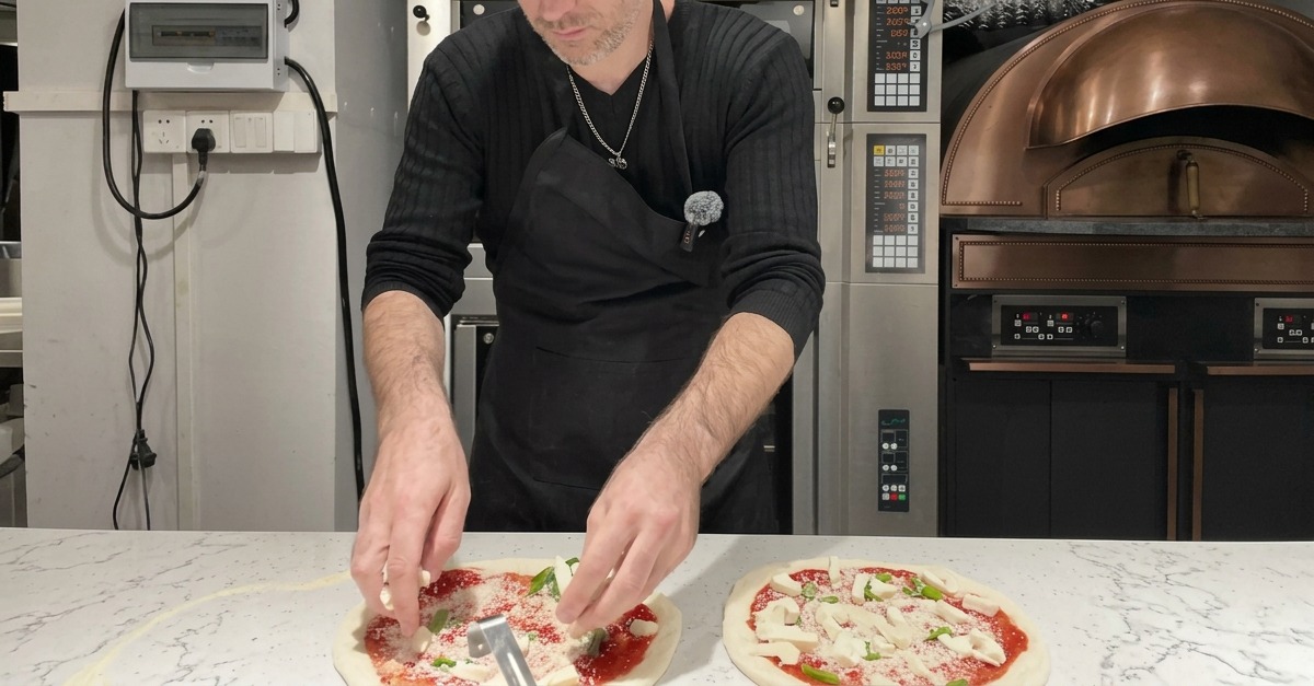 Chunks of white mozzarella cheese being scattered evenly across a pizza topped with sauce, basil, and Parmesan.
