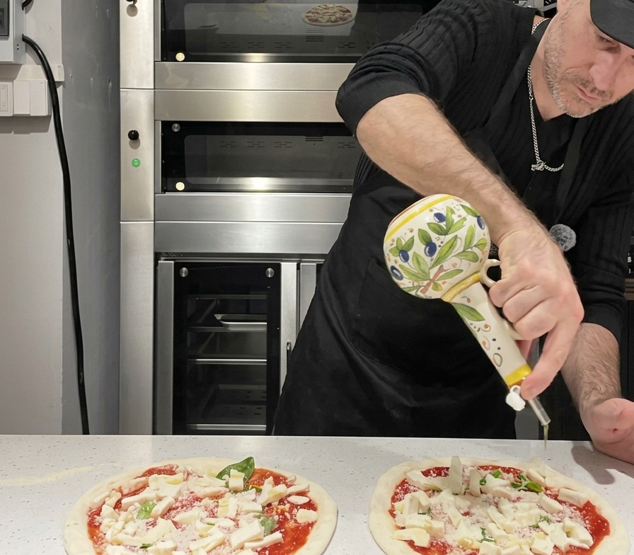 Olive oil being drizzled from a decorative ceramic cruet over an assembled raw Margherita pizza.