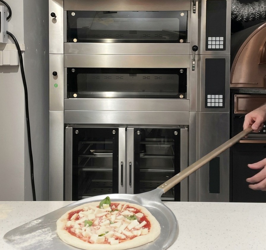 An assembled raw pizza on a metal pizza peel being slid into a commercial hot deck oven.
