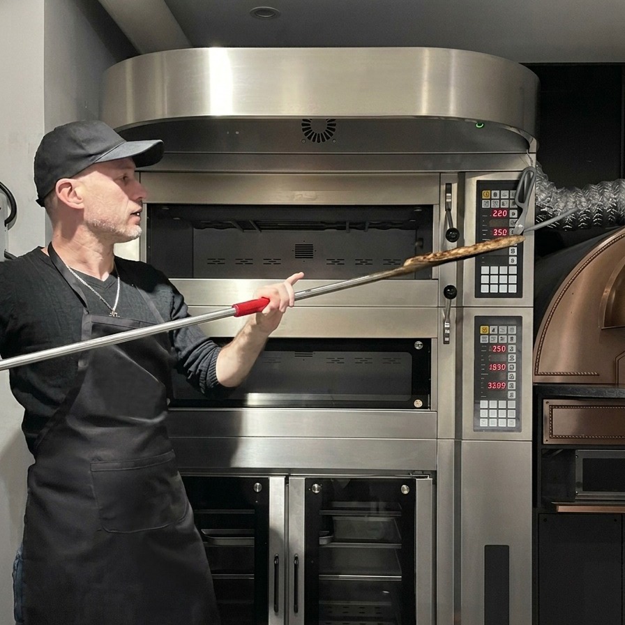 Chef using a long metal pizza peel to retrieve a baked pizza from a large professional oven.