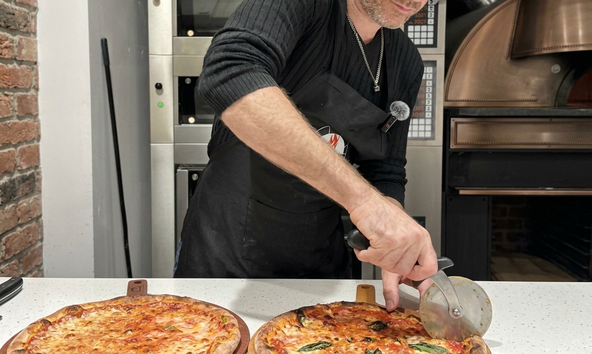 Chef slicing a fresh Margherita pizza on a wooden serving board using a round pizza cutter.