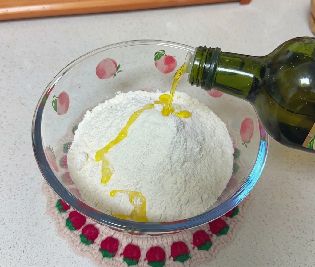 Olive oil is being poured from a dark green glass bottle onto a mound of white flour inside a glass bowl decorated with peach patterns.