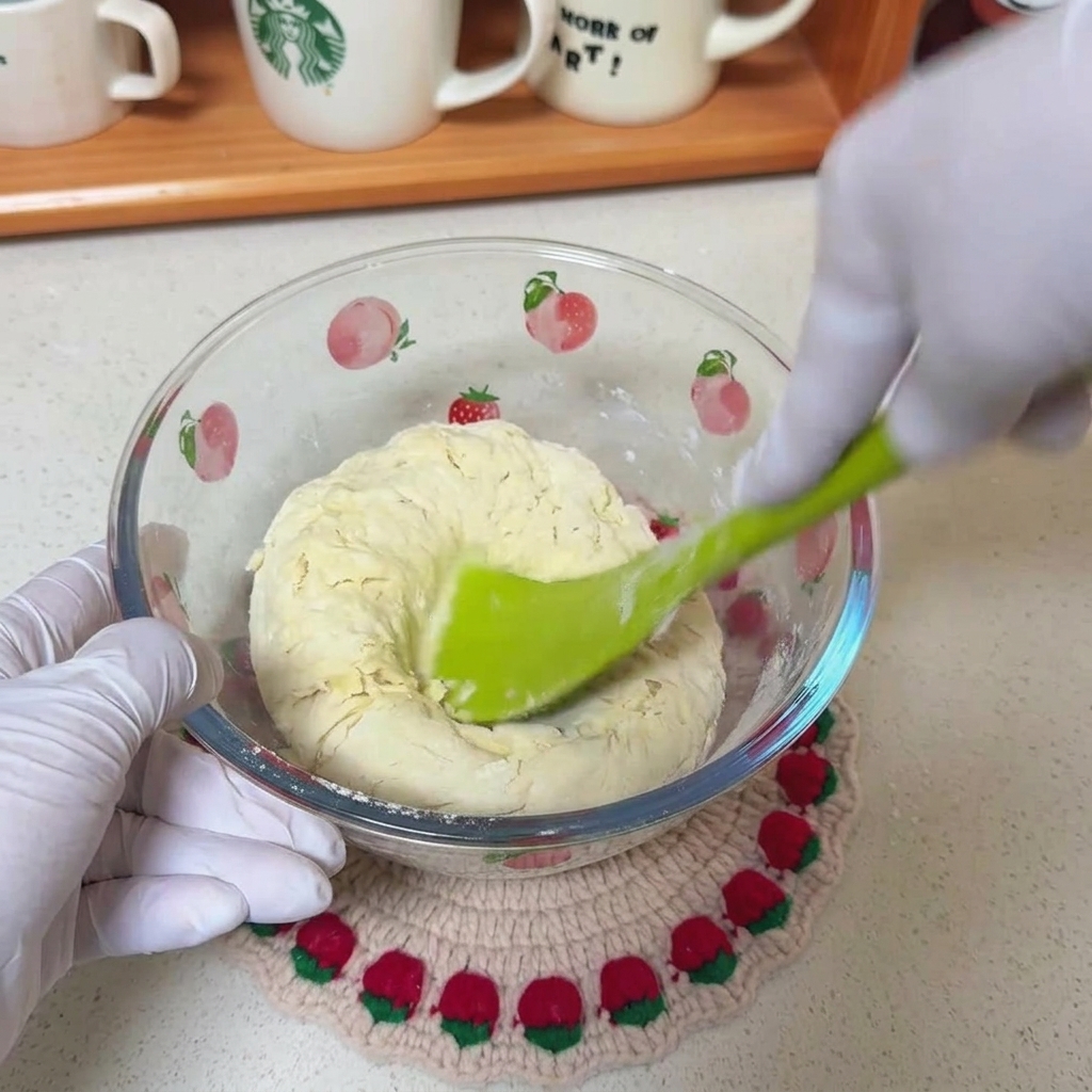A green spatula stirring a shaggy mixture of flour, water, and oil in a clear glass bowl decorated with fruit patterns.