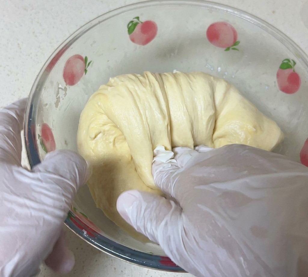 A person wearing white gloves stretching a smooth, pale yellow dough inside a glass bowl.