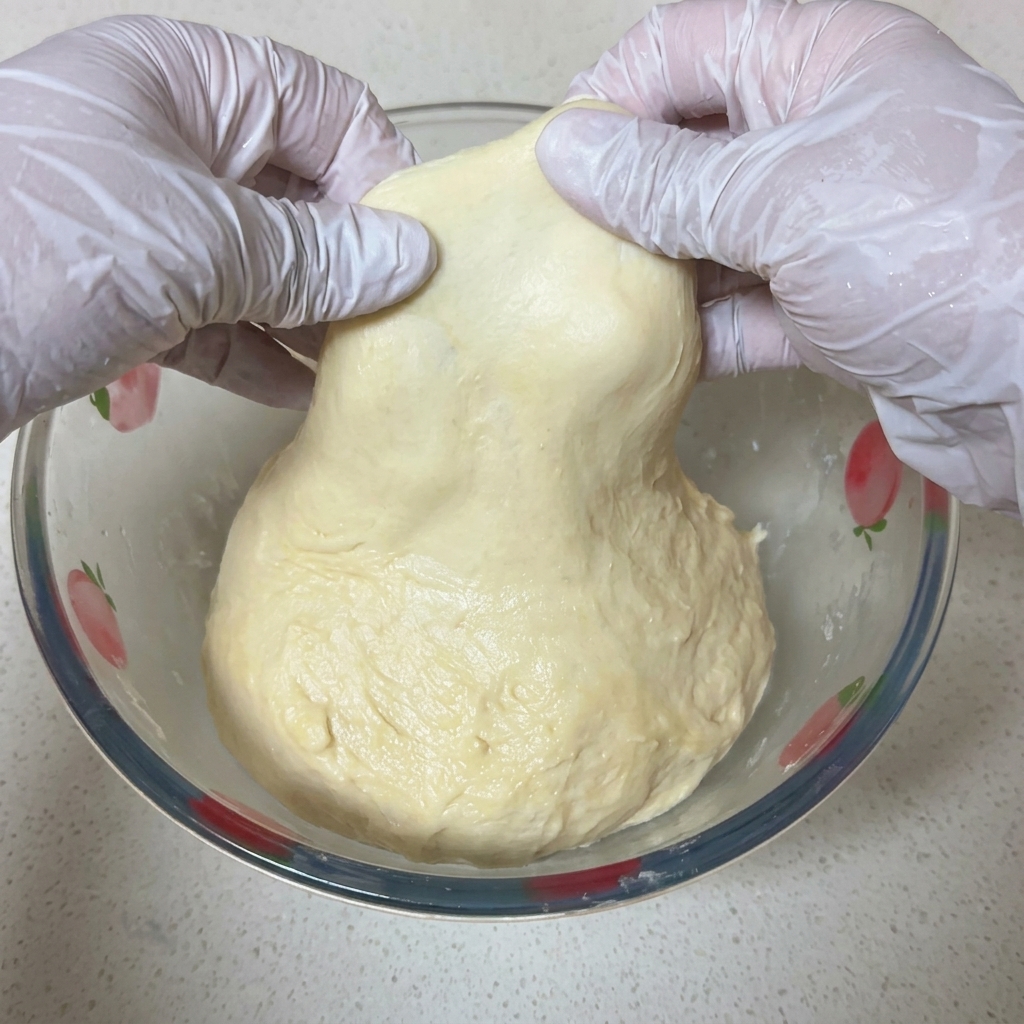 Two hands in white gloves carefully stretch a piece of pale pizza dough to show a thin, translucent membrane against the light.