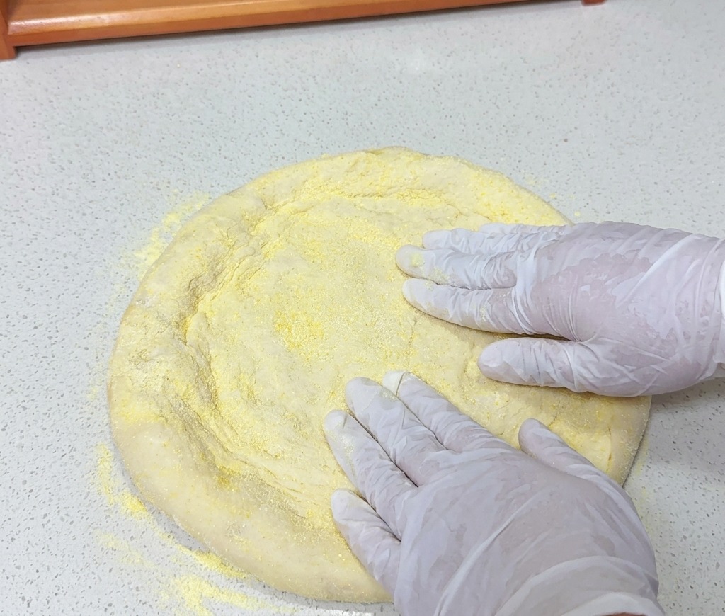 Hands in white gloves pressing down on a round piece of pizza dough covered in yellow cornmeal on a light countertop.