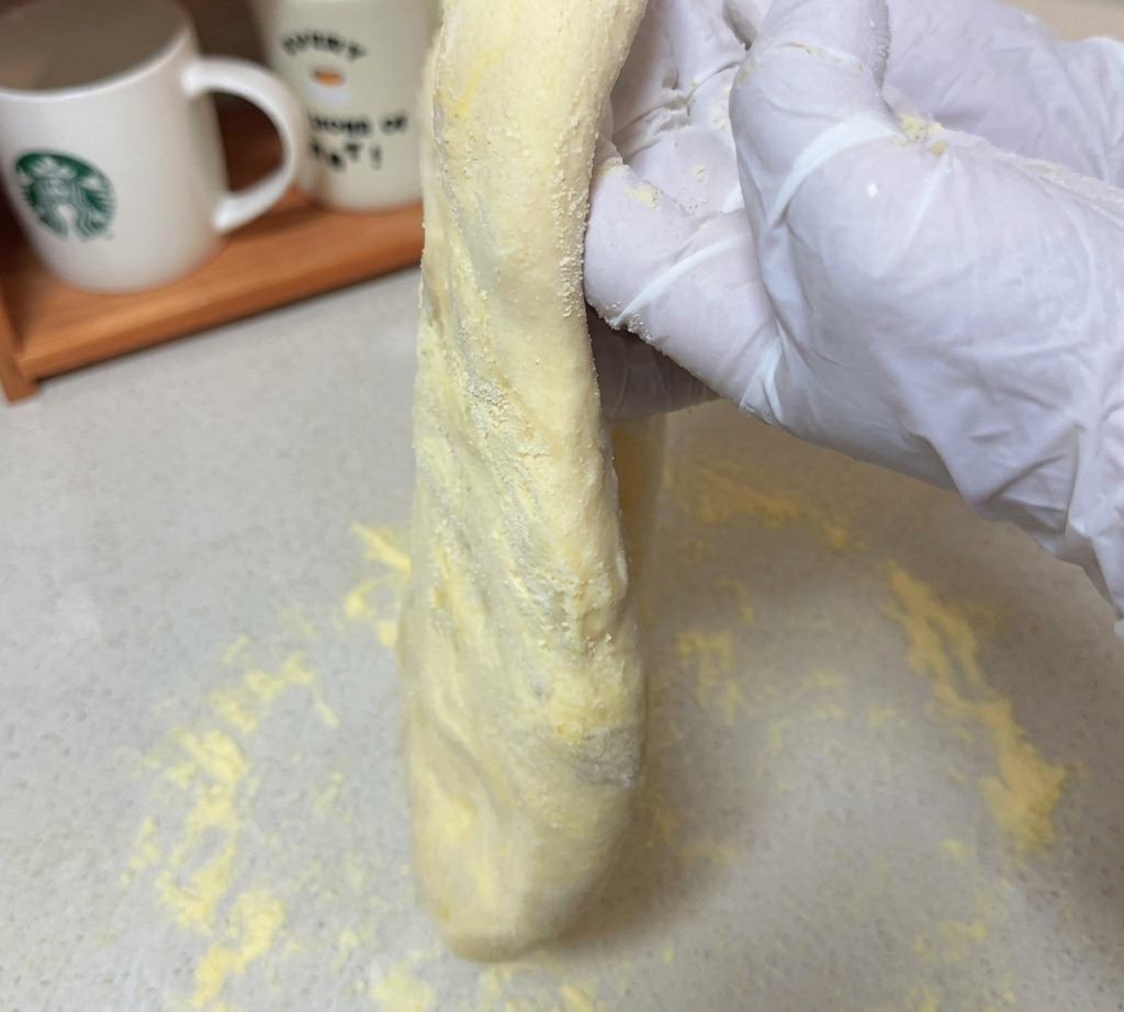 A long, stretched piece of raw pizza dough being held up by gloved hands over a floured surface.