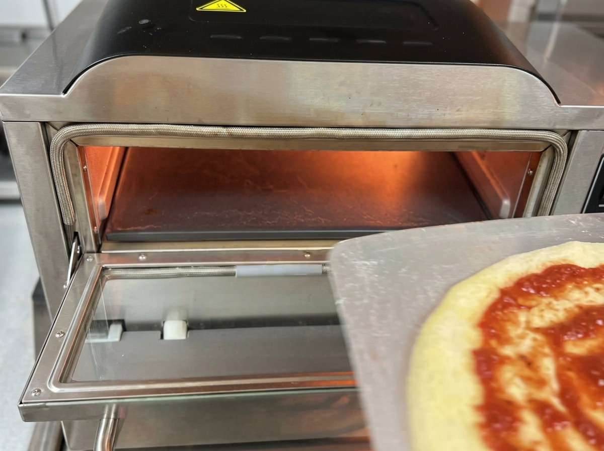 A raw pizza topped with tomato sauce and mozzarella being transferred from a metal pizza peel into the glowing interior of a professional electric pizza oven.