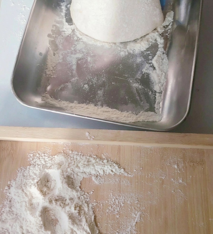 A round ball of pale pizza dough being lifted from a stainless steel tray onto a wooden board sprinkled with flour.