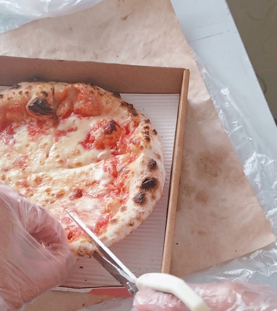 A person wearing plastic gloves using white kitchen scissors to cut a hot cheese pizza into wedges inside a cardboard delivery box.