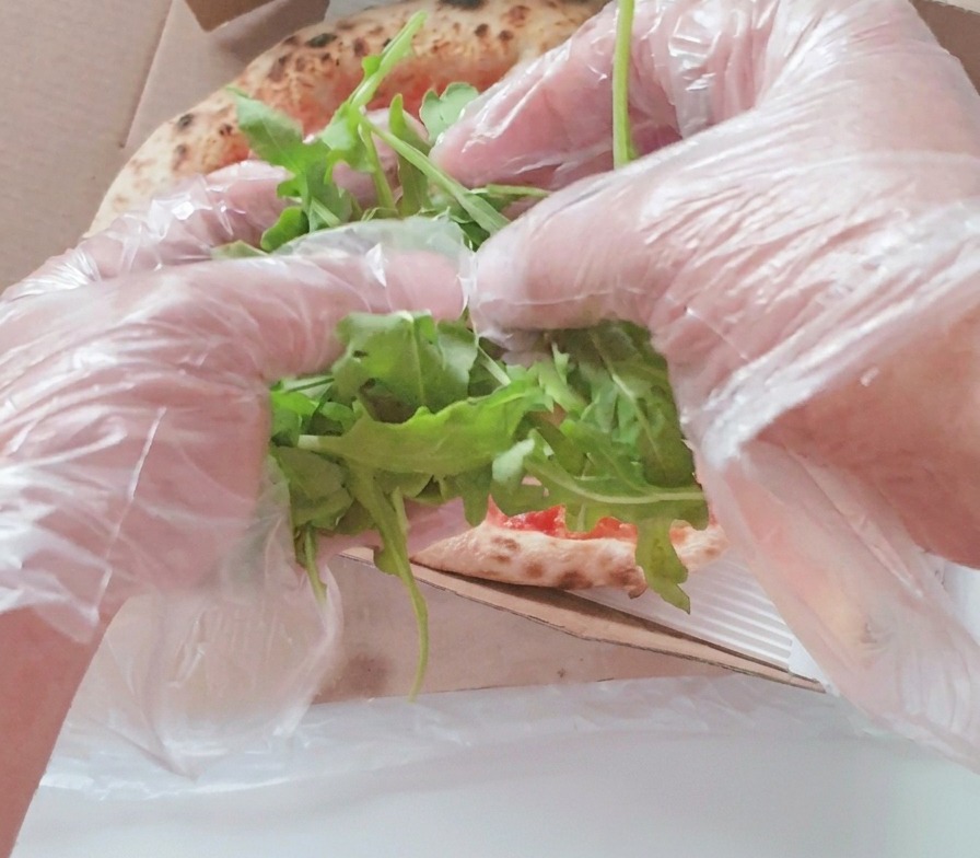 Hands in clear plastic gloves placing a heap of fresh green arugula leaves onto the center of a baked pizza in a box.