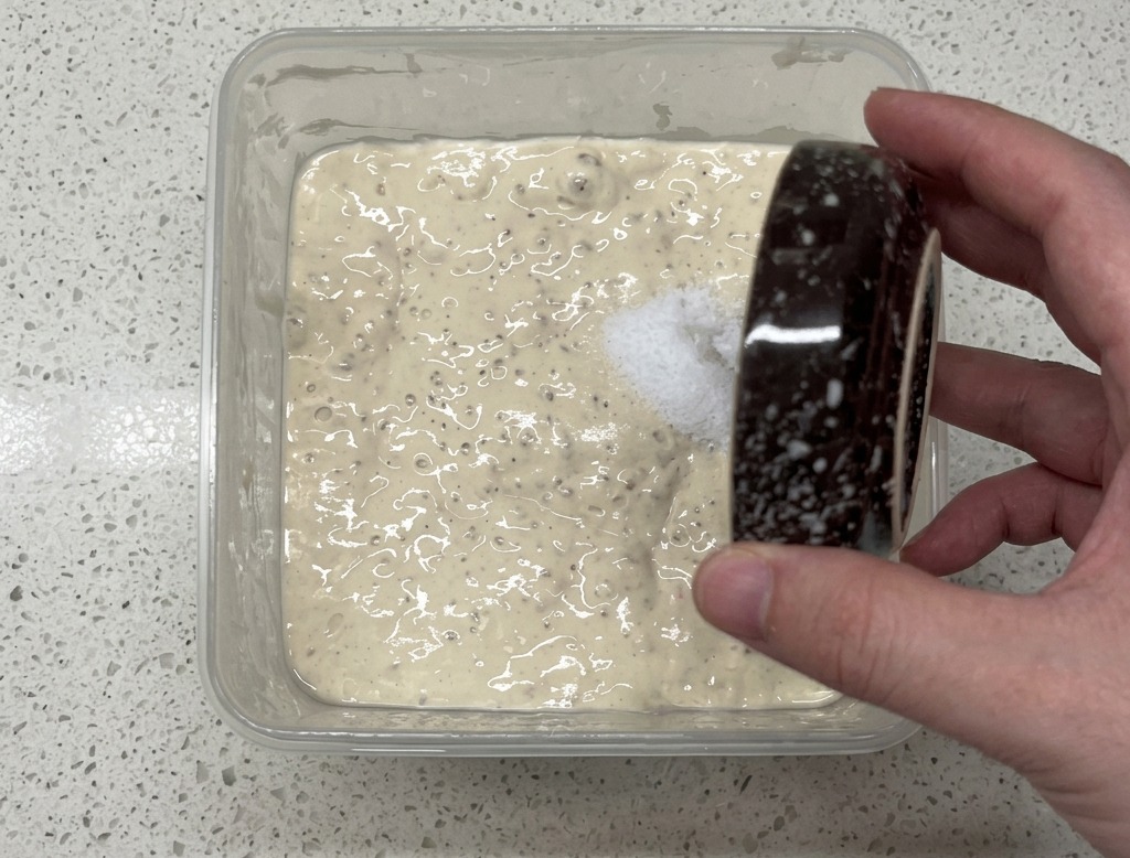 A hand pouring salt from a small speckled bowl into a square plastic container containing a bubbly, pale poolish starter.