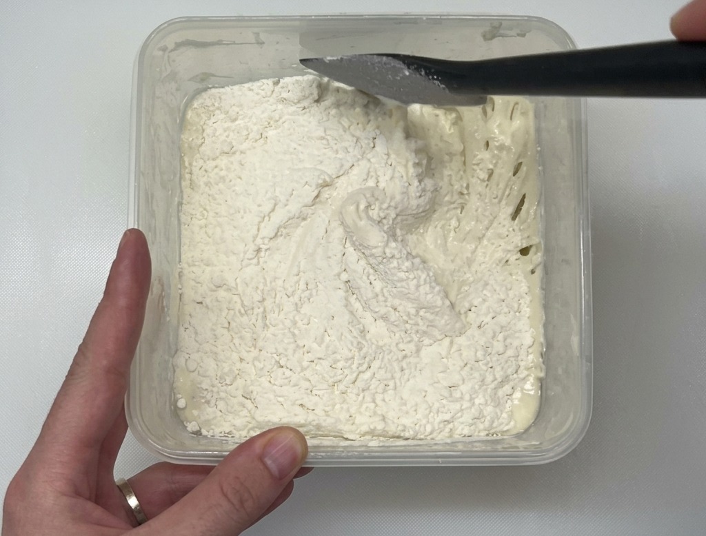 A black spatula mixing white flour into a wet dough inside a clear plastic container on a white countertop.