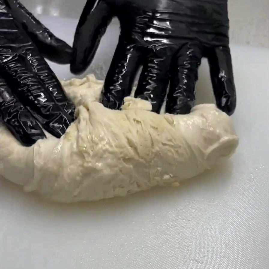A person wearing black gloves stretching and folding a piece of pale, wet dough on a white surface.