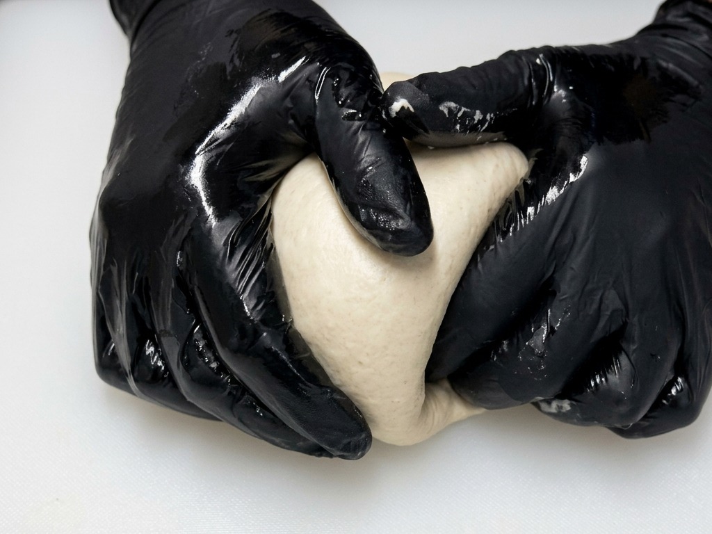Hands in black gloves shaping a smooth, round ball of pizza dough by dragging it across a white board.