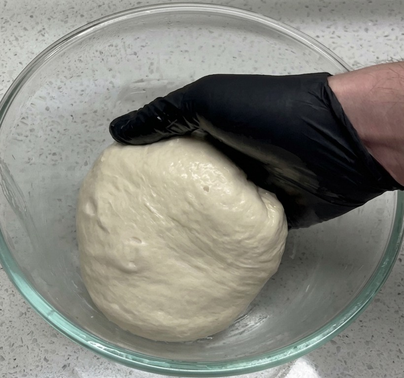 A hand in a black glove placing a smooth dough ball into a large, clear glass bowl on a granite-style countertop.