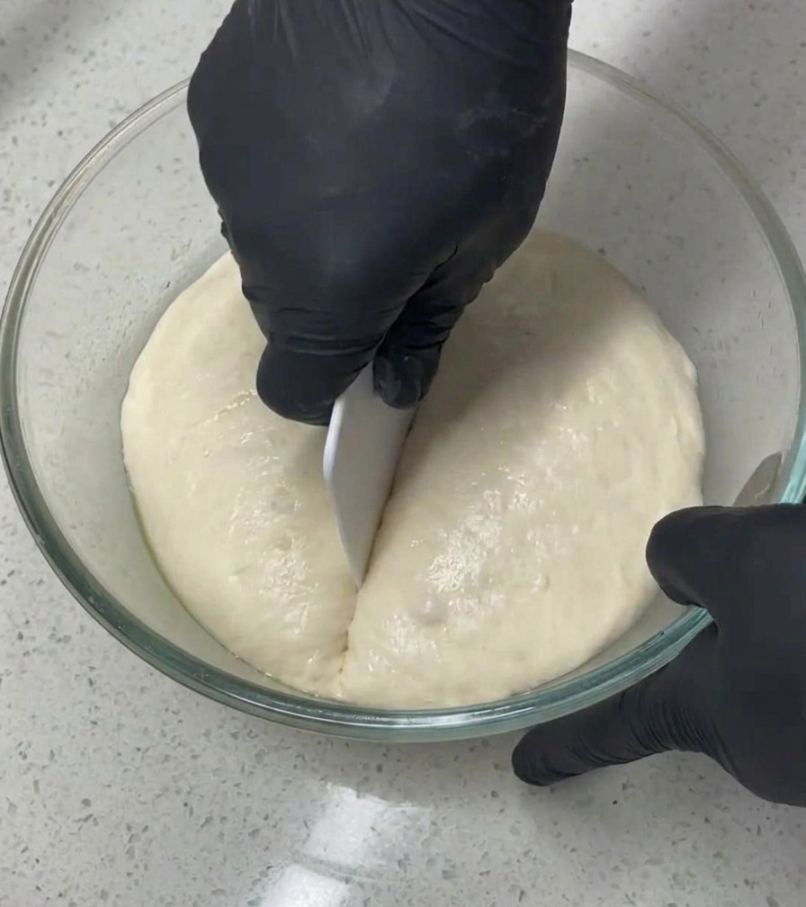 A person wearing black gloves uses a white bench scraper to cut a large, airy mass of dough in half inside a glass bowl.