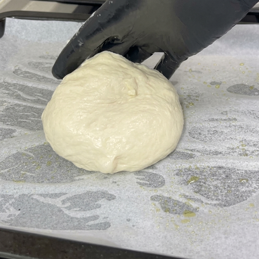 A gloved hand places a soft, rounded ball of pizza dough onto a tray lined with white parchment paper.