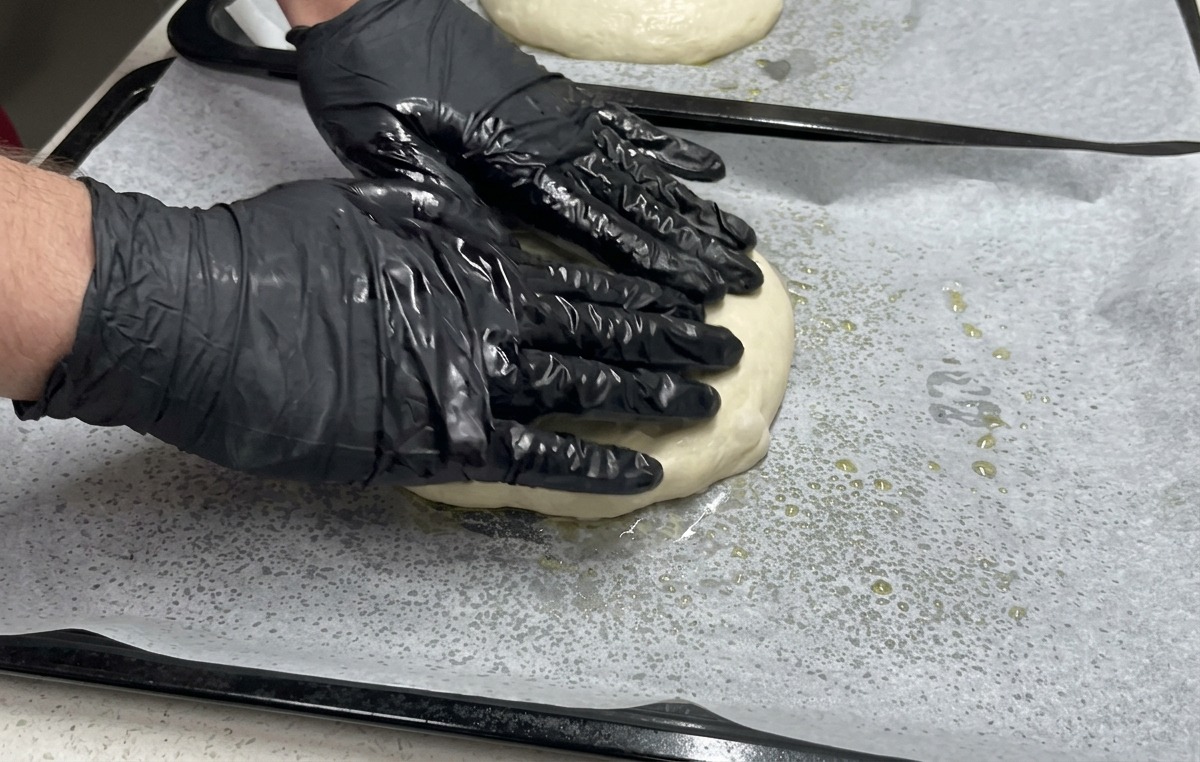 Two hands in black gloves press down on a flattened piece of dough on a parchment-lined baking sheet to spread it to the edges.
