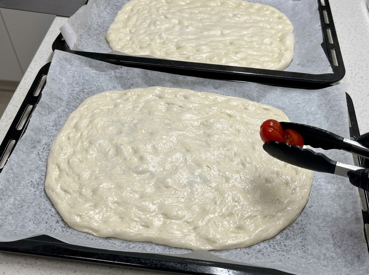 Using black kitchen tongs, a halved cherry tomato is placed onto a large sheet of flattened raw pizza dough.