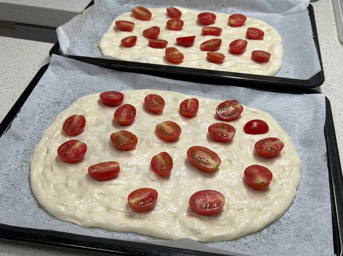 Two baking trays shown with flattened pizza dough topped with numerous halved cherry tomatoes, ready for seasoning.