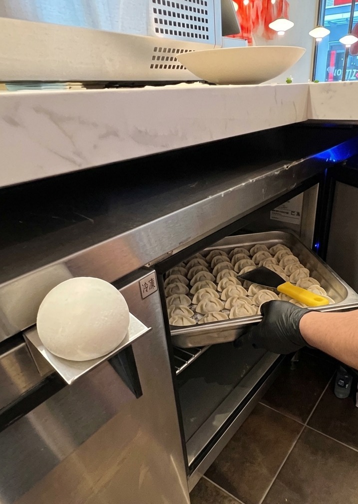 A gloved hand pulling a stainless steel tray filled with round, white portions of pizza dough from a professional cooling unit.