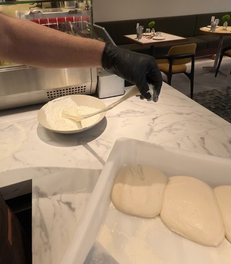 A gloved hand lifting a stretchy piece of fermented pizza dough out of a bowl of white flour.