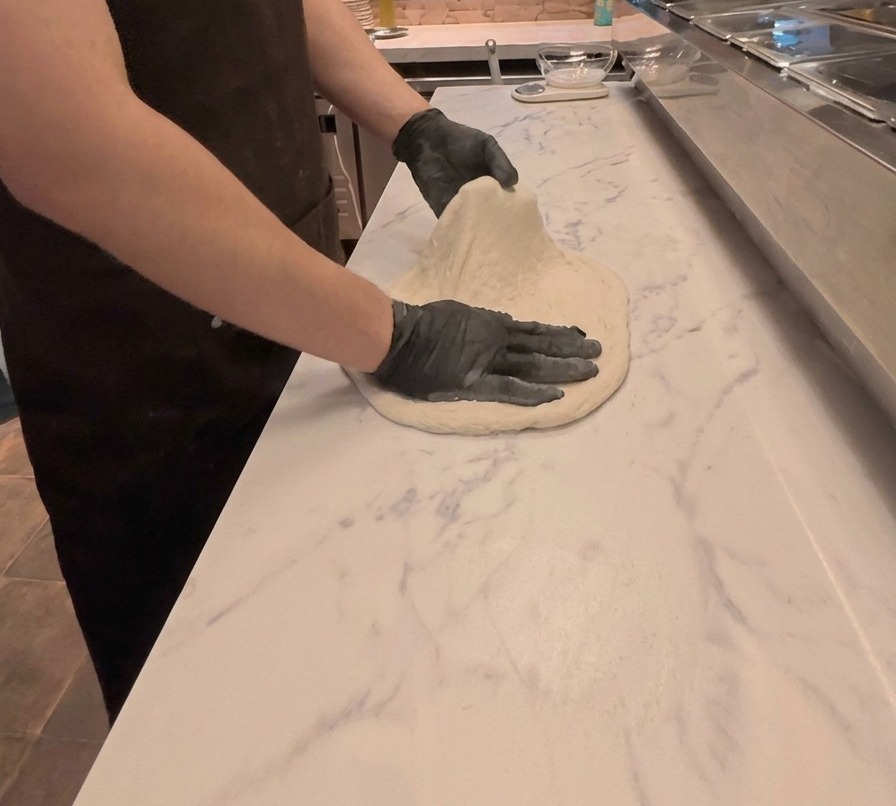 A cook using both hands to stretch and flatten pizza dough on a white marble-patterned countertop.