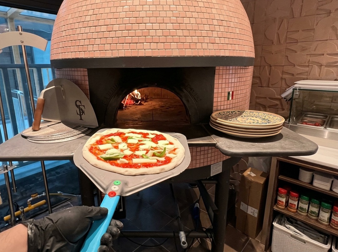 An unbaked pizza topped with tomato sauce and mozzarella sitting on a metal peel at the entrance of a pink-tiled wood-fired oven.