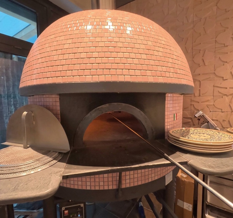 The exterior of a pink-tiled dome pizza oven with a pizza visible baking inside near the glowing embers.