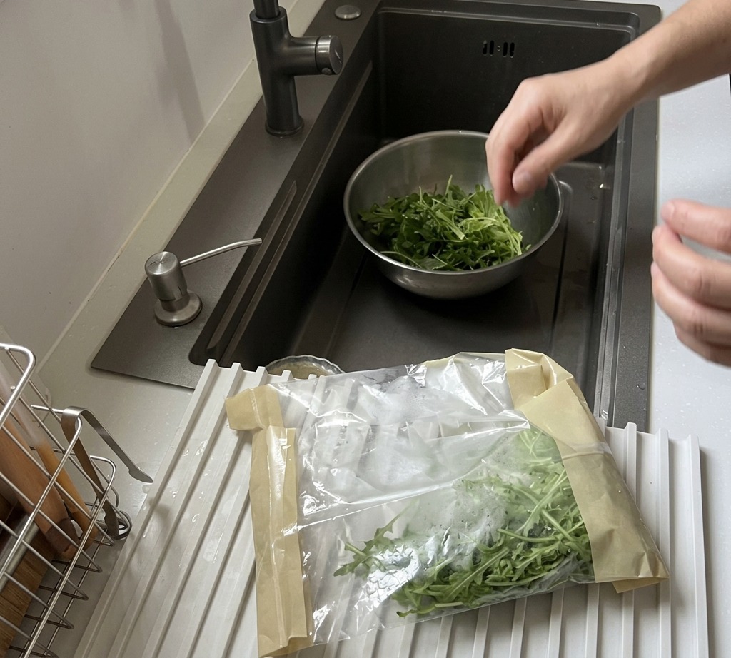 Fresh arugula leaves being washed in a stainless steel bowl under a kitchen faucet.