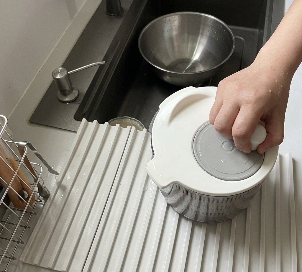 A person using a white manual salad spinner to dry fresh arugula leaves.
