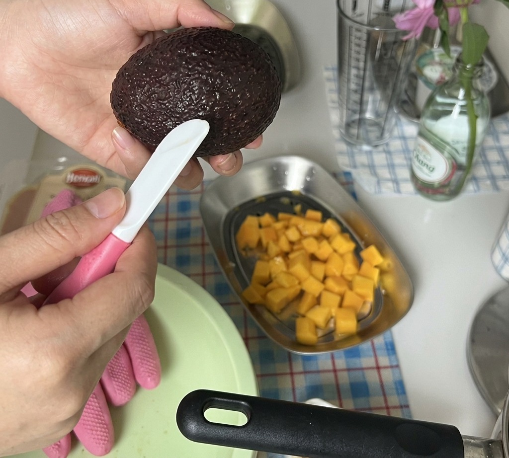 Hands slicing a peeled avocado half into small cubes over a tray of diced mango