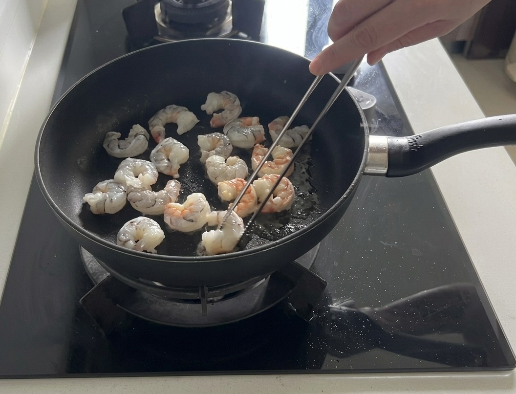Fresh shrimp being sautéed in a dark frying pan with olive oil, with some turning pink and opaque.
