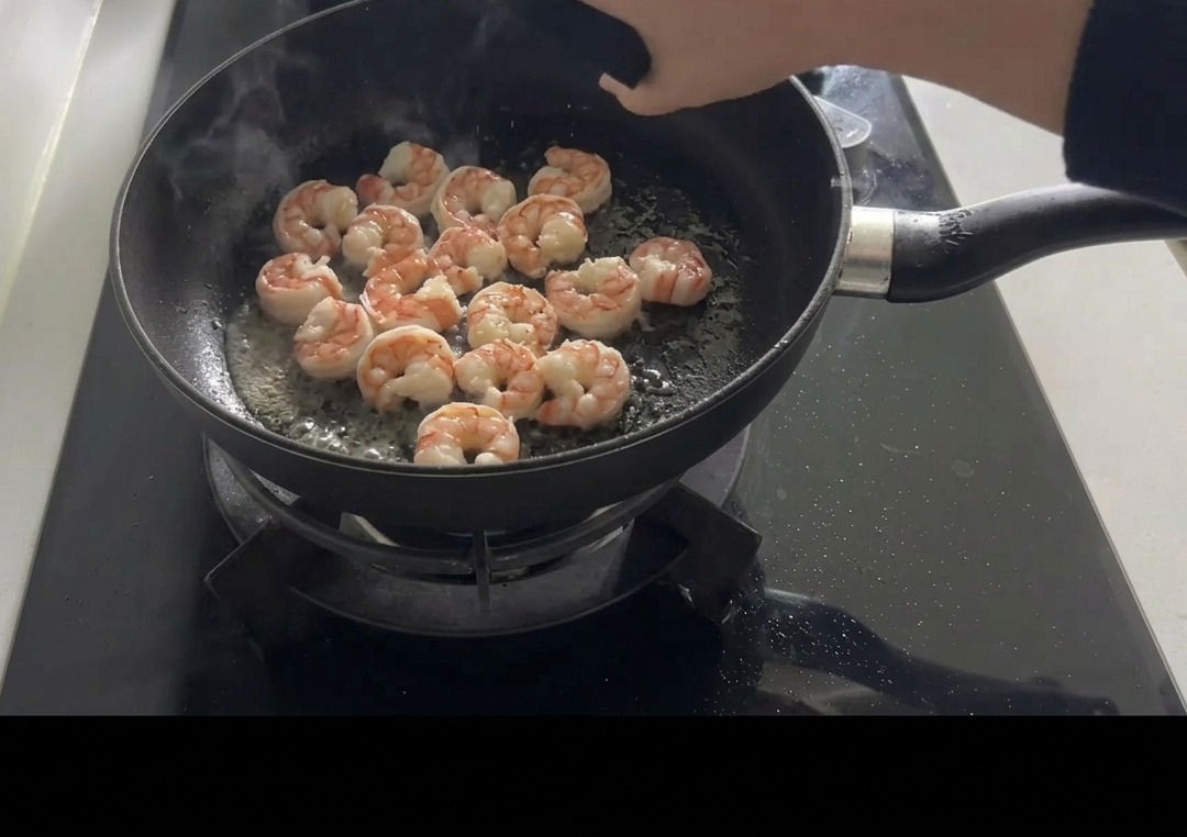 Sautéed pink shrimp in a black frying pan being seasoned with salt and pepper by hand.