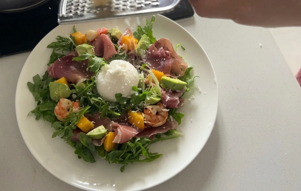 A plated arugula salad topped with a large ball of white burrata cheese and thin ribbons of cured prosciutto.